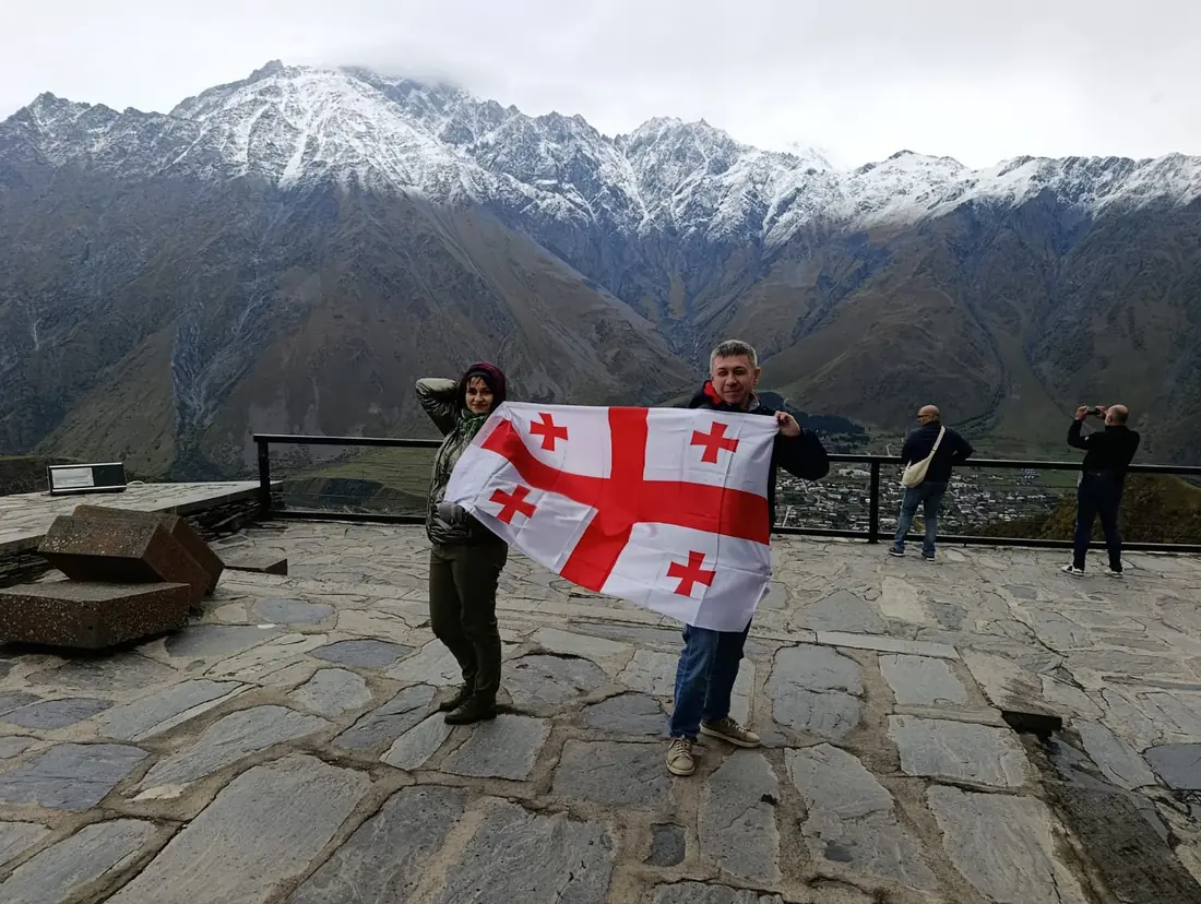 Travellers with the Georgian flag on the Gergeti church terrace, Stepantsminda below